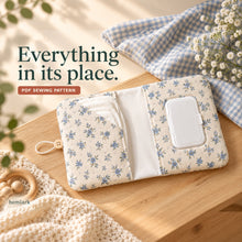 Open blue floral diaper wallet on a wooden table shows a plain diaper pocket, white wipes lid in its access window and a pale center lining.