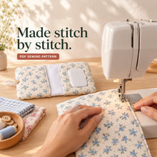 Hands guide a blue floral pocket panel beneath a sewing-machine presser foot, with a completed open diaper wallet behind it.