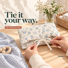 Two hands tie the ivory ribbon closure of a blue floral diaper wallet on a sunlit wooden sewing table.