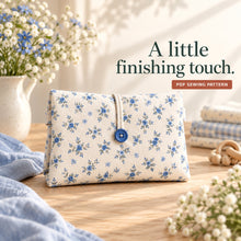 Blue floral cotton diaper clutch with one blue button and a thin ivory elastic loop, beside gingham fabric and flowers.