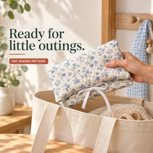 A hand holds a blue floral diaper clutch with ivory ties above a canvas tote containing a soft blanket.