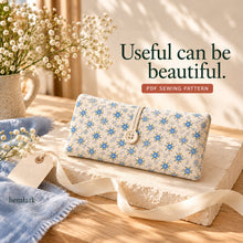 A closed blue floral diaper clutch with a wooden button and slim ivory loop rests on a curved paper plinth beside a gift tag and loose ribbon.
