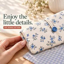 A hand holds the rounded, neatly topstitched corner of a blue floral diaper clutch with a blue button and narrow ivory loop.